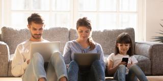 Vater, Mutter und Kind sitzen vor einer Couch auf dem Boden, jeweils mit einem technischen Gerät in der Hand und schauen darauf. 